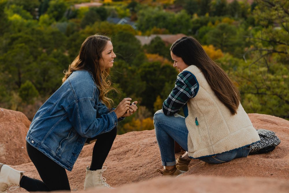 A surprise proposal moment unfolds on red rocks with mountains and forest in the background.