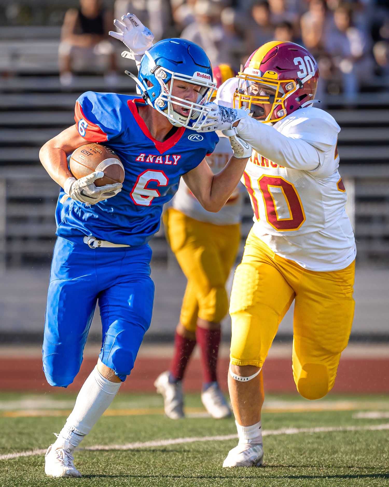 High school football player in blue jersey carries ball while being tackled by defender in yellow uniform during game action.