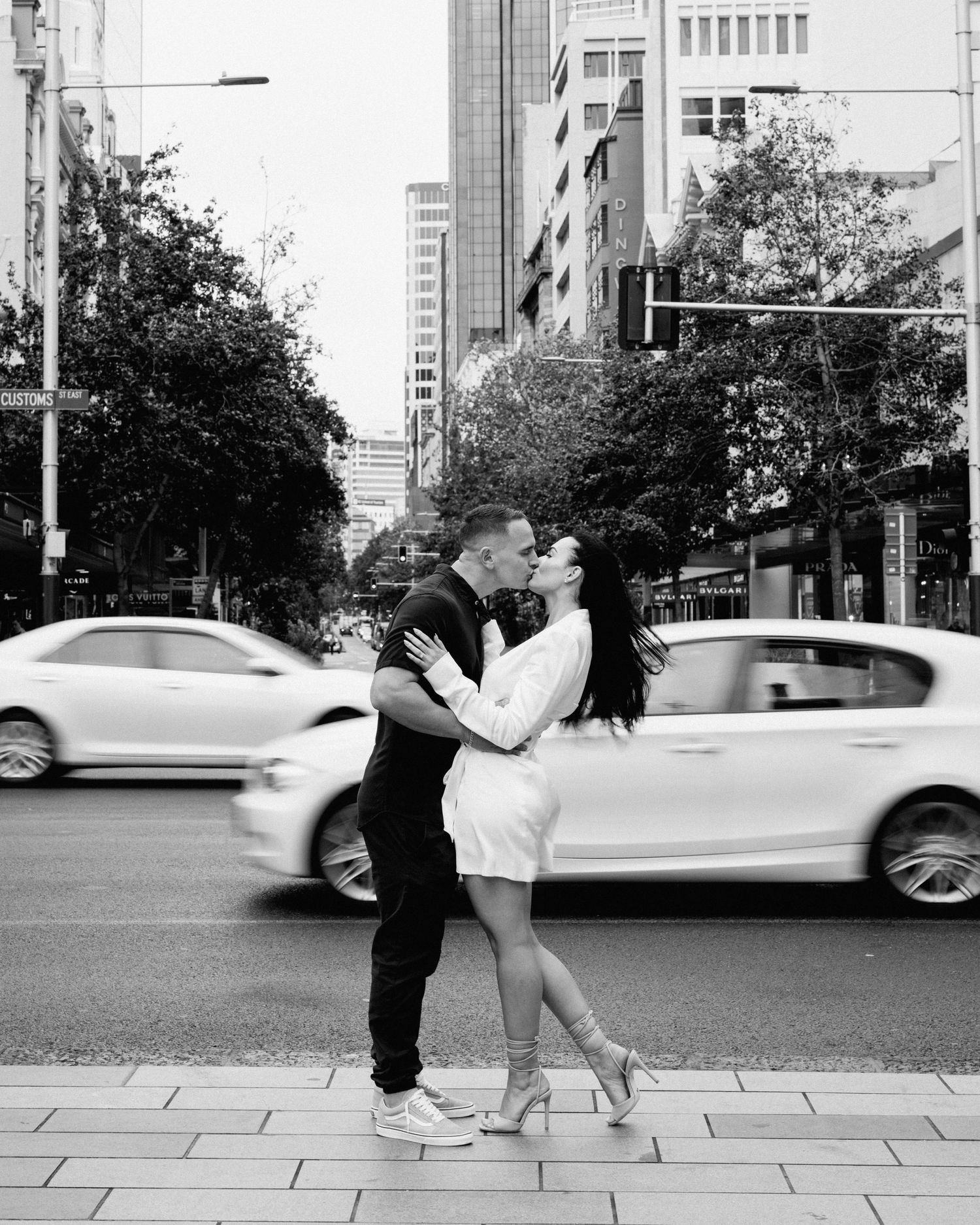 Romantic couple embraces on city sidewalk as cars blur past in black and white urban scene.