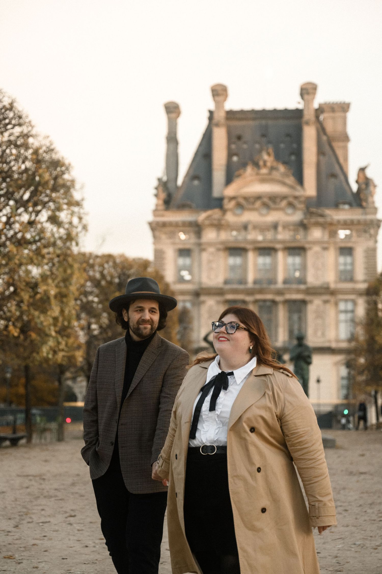 Stylishly dressed pair stands in front of historic Parisian architecture at the Louvre.