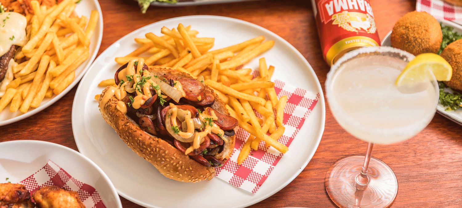 Gourmet hot dog with toppings served with french fries and a cocktail on red checkered placemat.