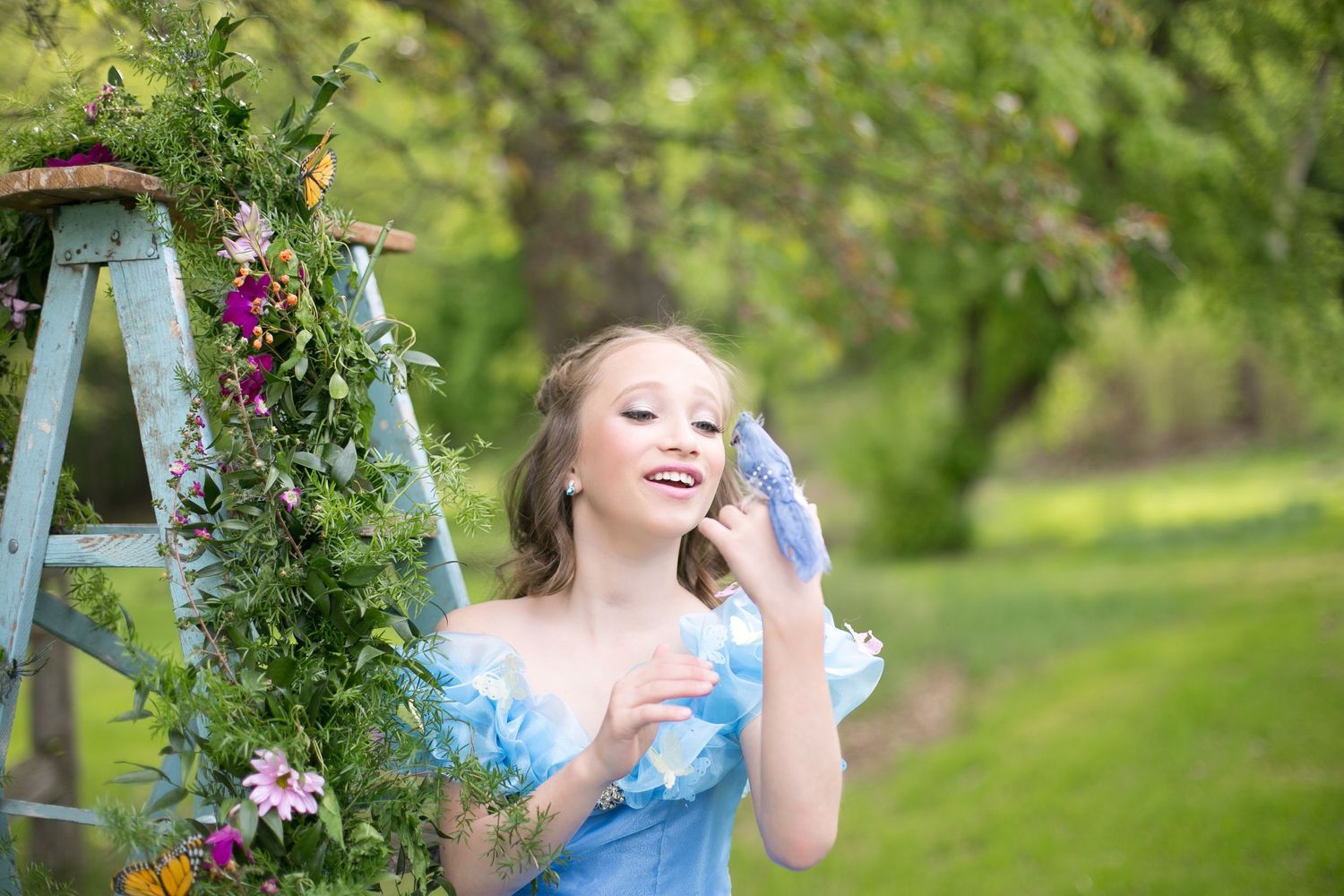 Young blonde girl in blue princess dress holds blue bird near flower-adorned ladder in lush green garden.