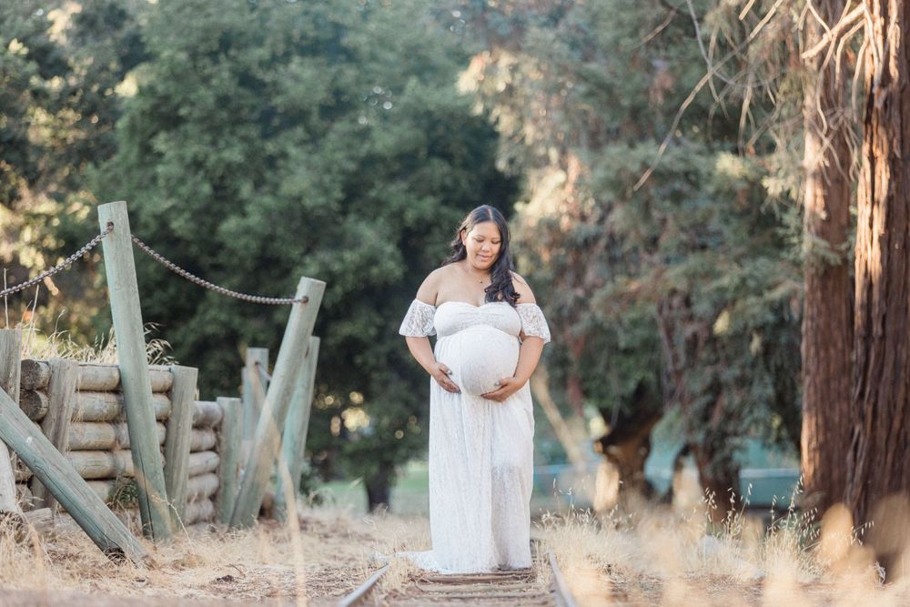 A maternity photo of a person in a flowing white dress standing on a rustic path surrounded by trees at sunset.