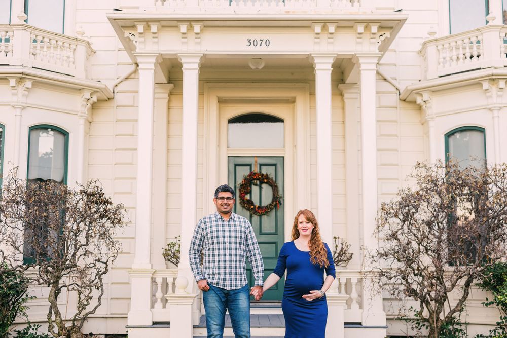 A couple stands in front of a grand white Victorian mansion with pillars.