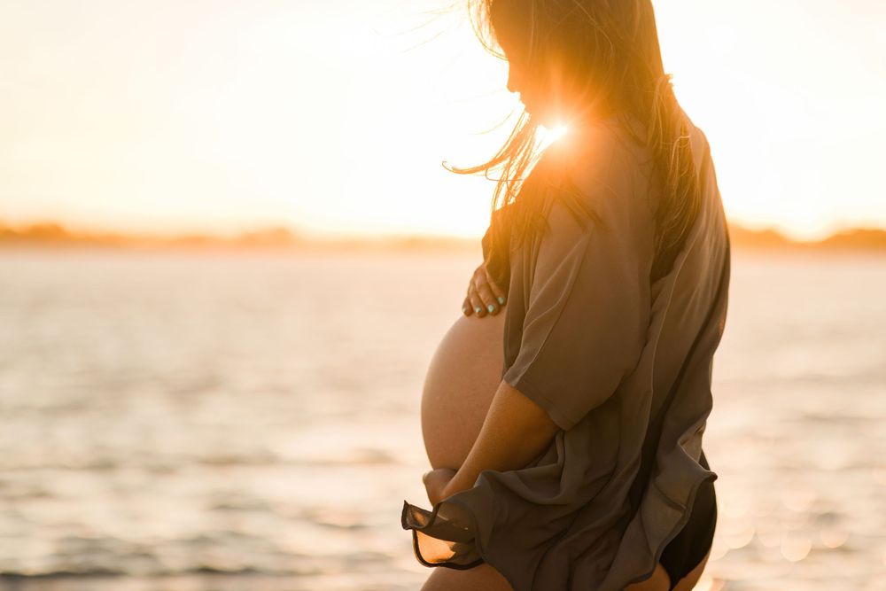 Silhouette of a pregnant woman standing by the water during sunset.