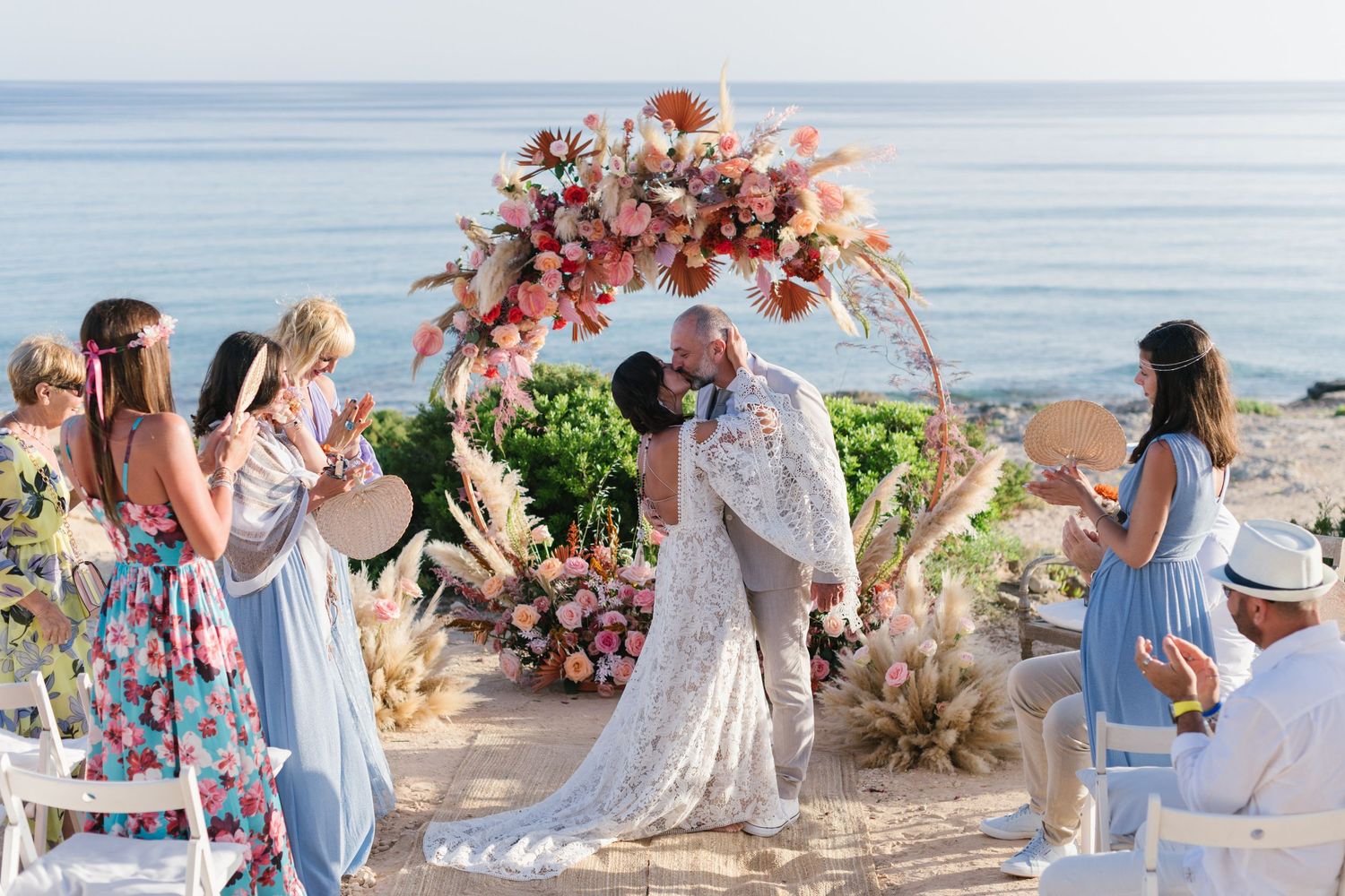 A bride and groom kiss under an arch of flowers with a background of blue sea in Formentera