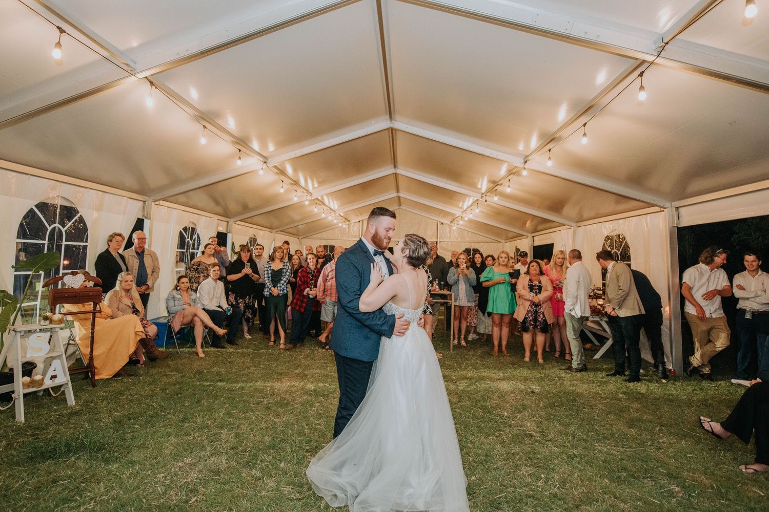 A couple shares their first dance surrounded by guests in an elegantly lit reception tent.