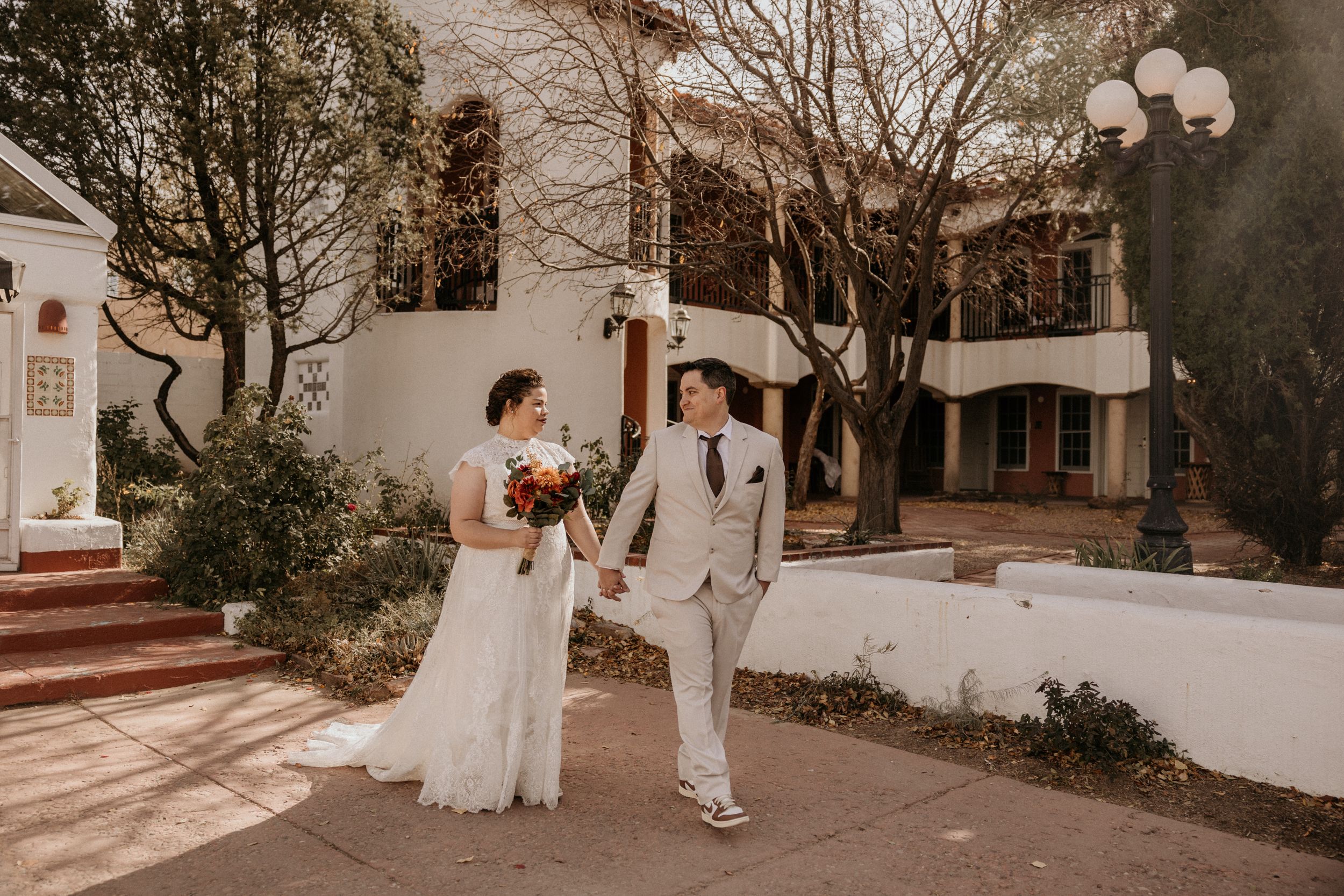 Bride and groom hold hands and smile at each other outside a white stucco wedding venue.