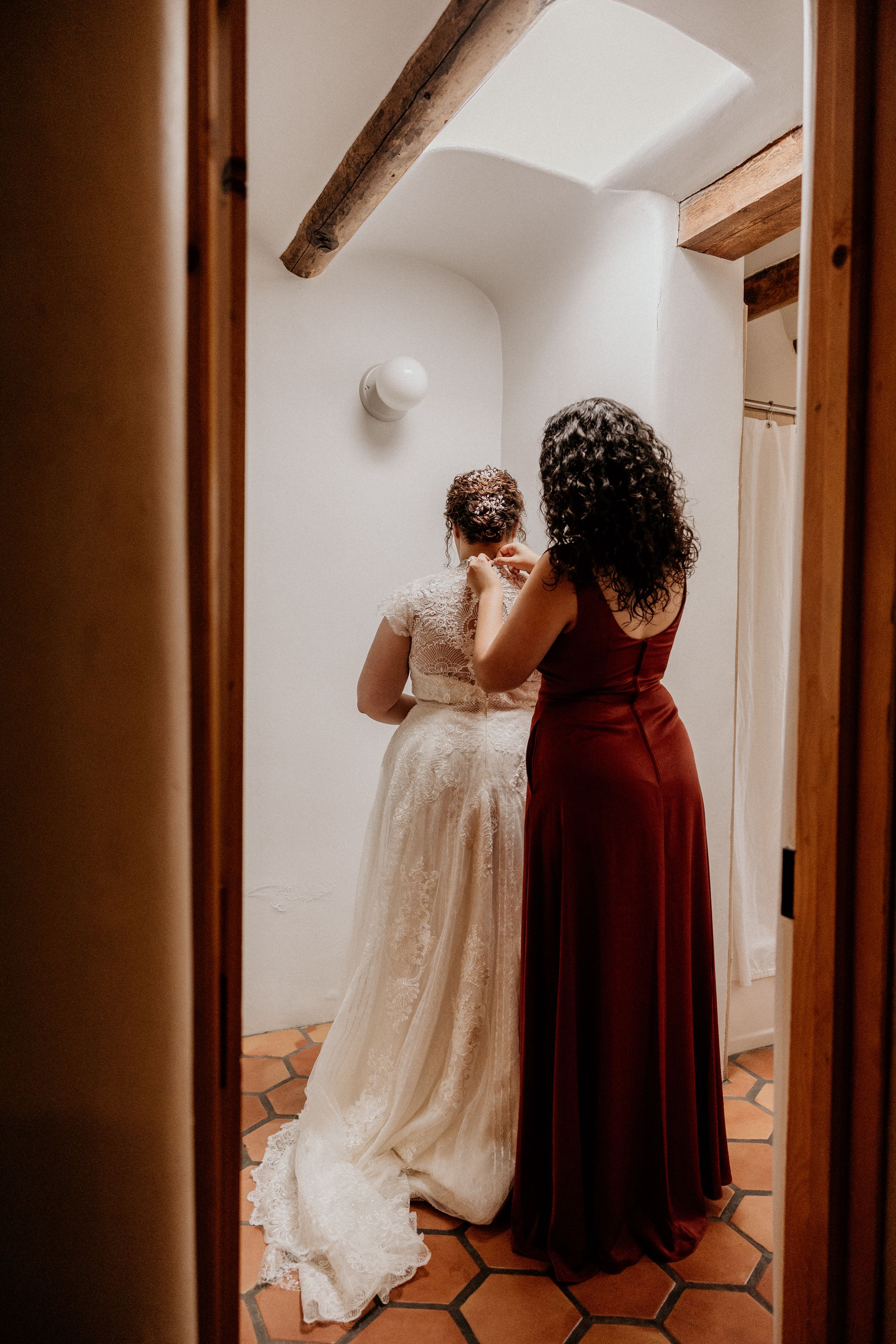 Bride in lace gown gets help from bridesmaid in burgundy dress in rustic adobe venue.