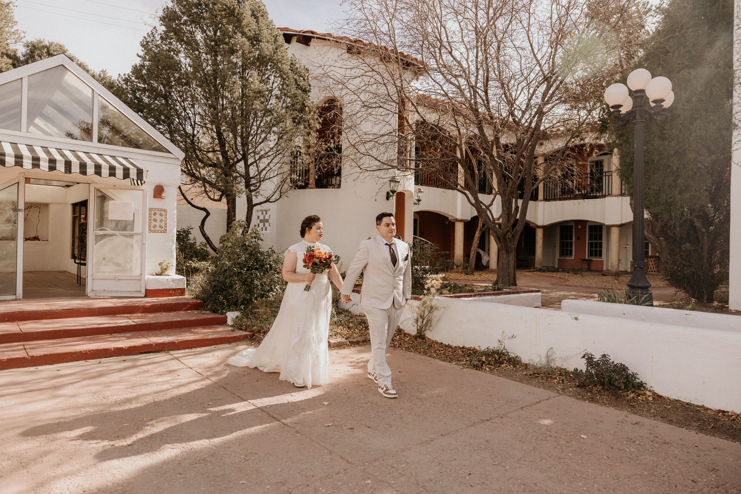 Wedding couple walks hand-in-hand outside a white Spanish-style venue surrounded by trees.
