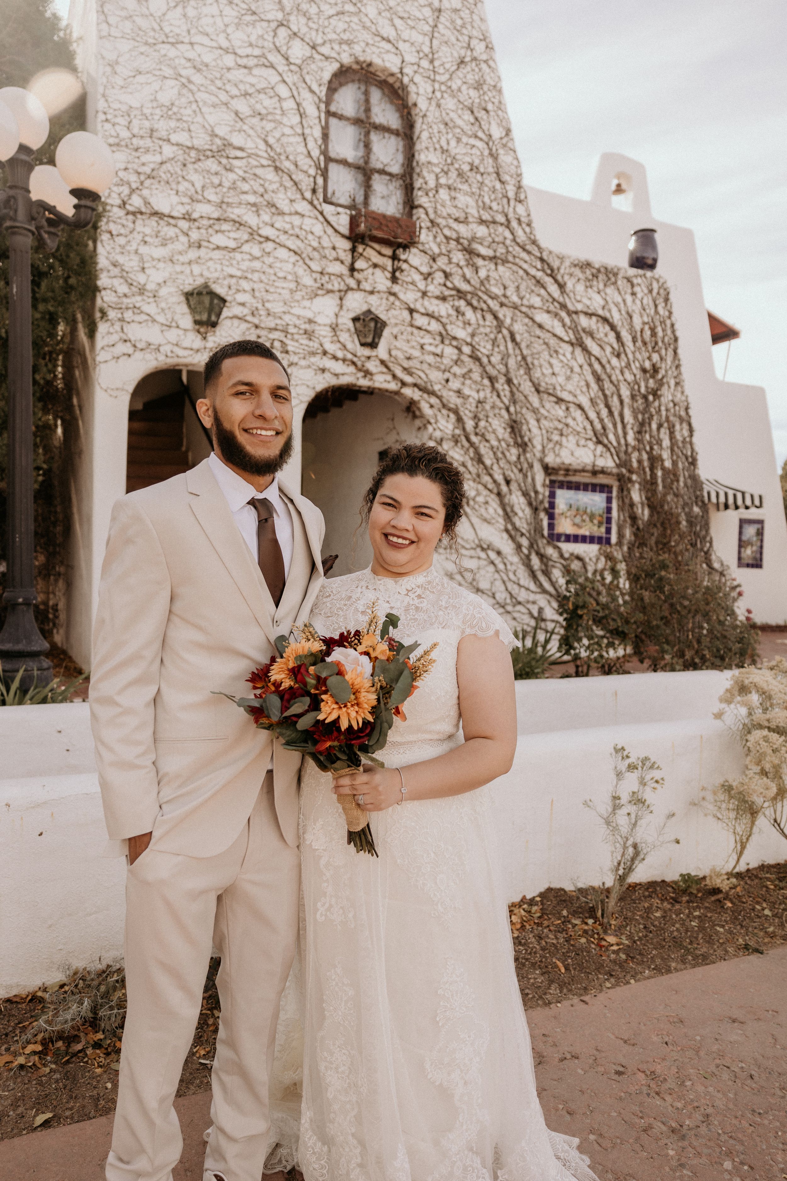 Smiling bride and groom pose together holding fall bouquet outside white adobe-style building.