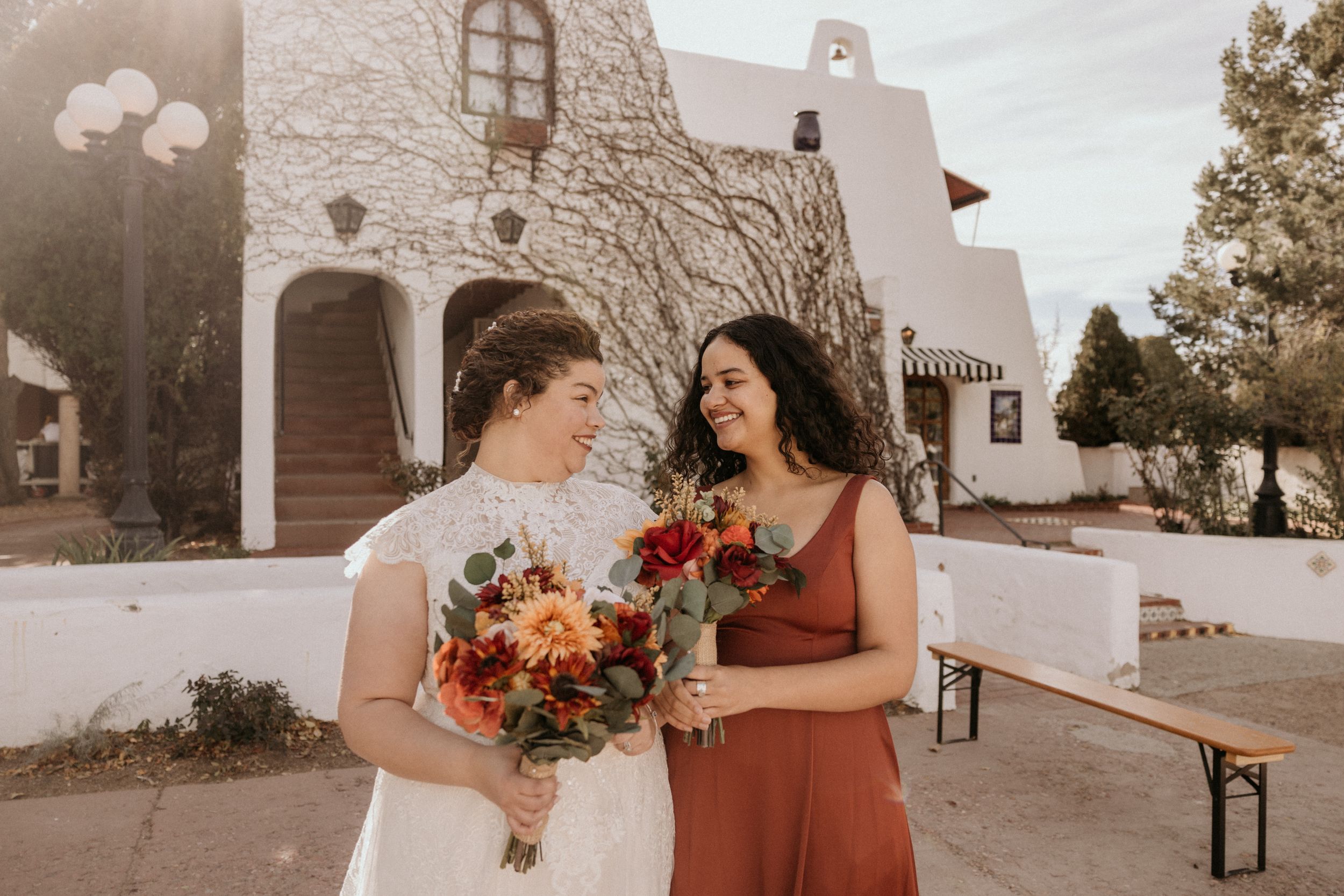 Bride and bridesmaid in terracotta dress share a joyful moment outside a white stucco building.