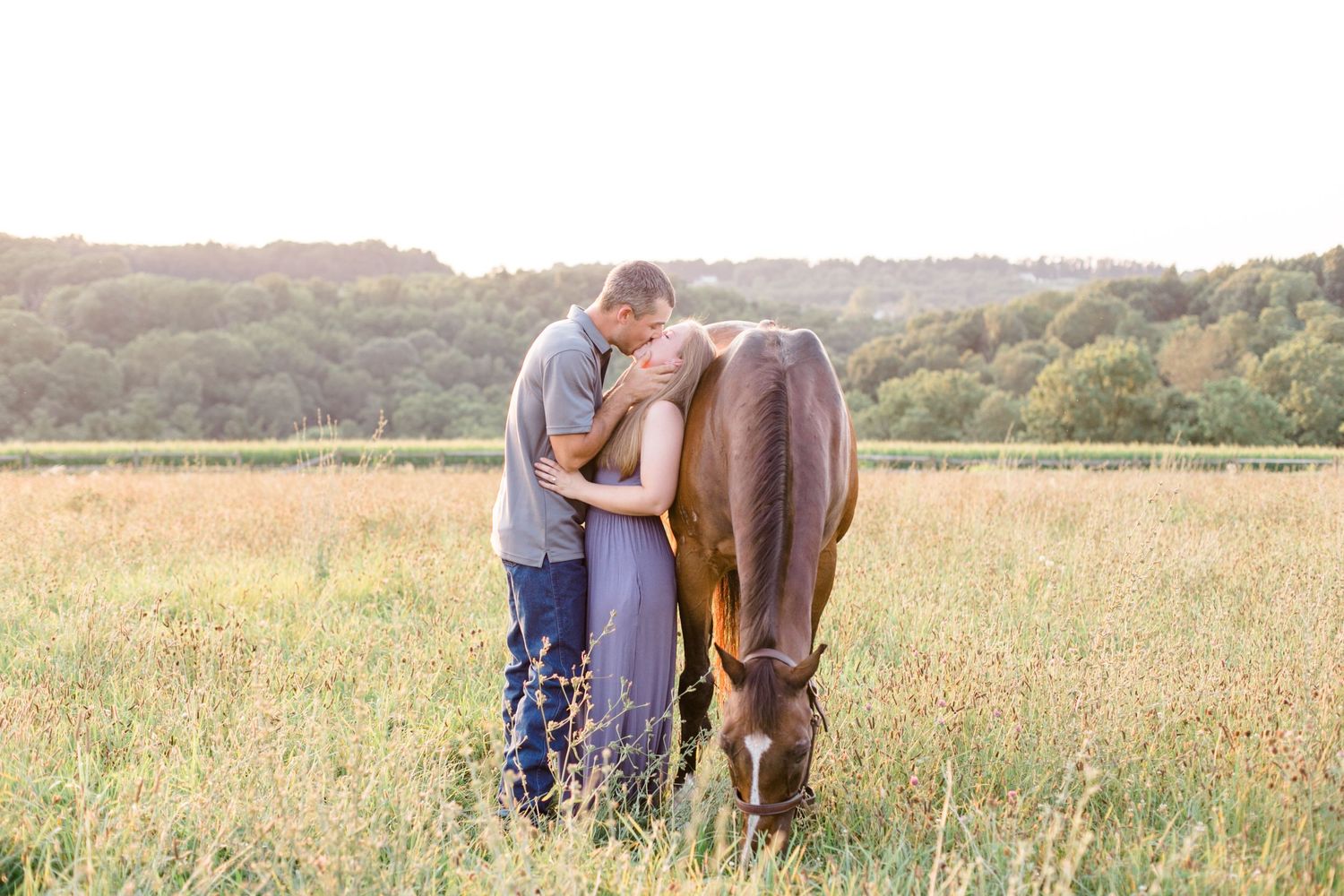 Couple stands with grazing horse in golden sunset field.