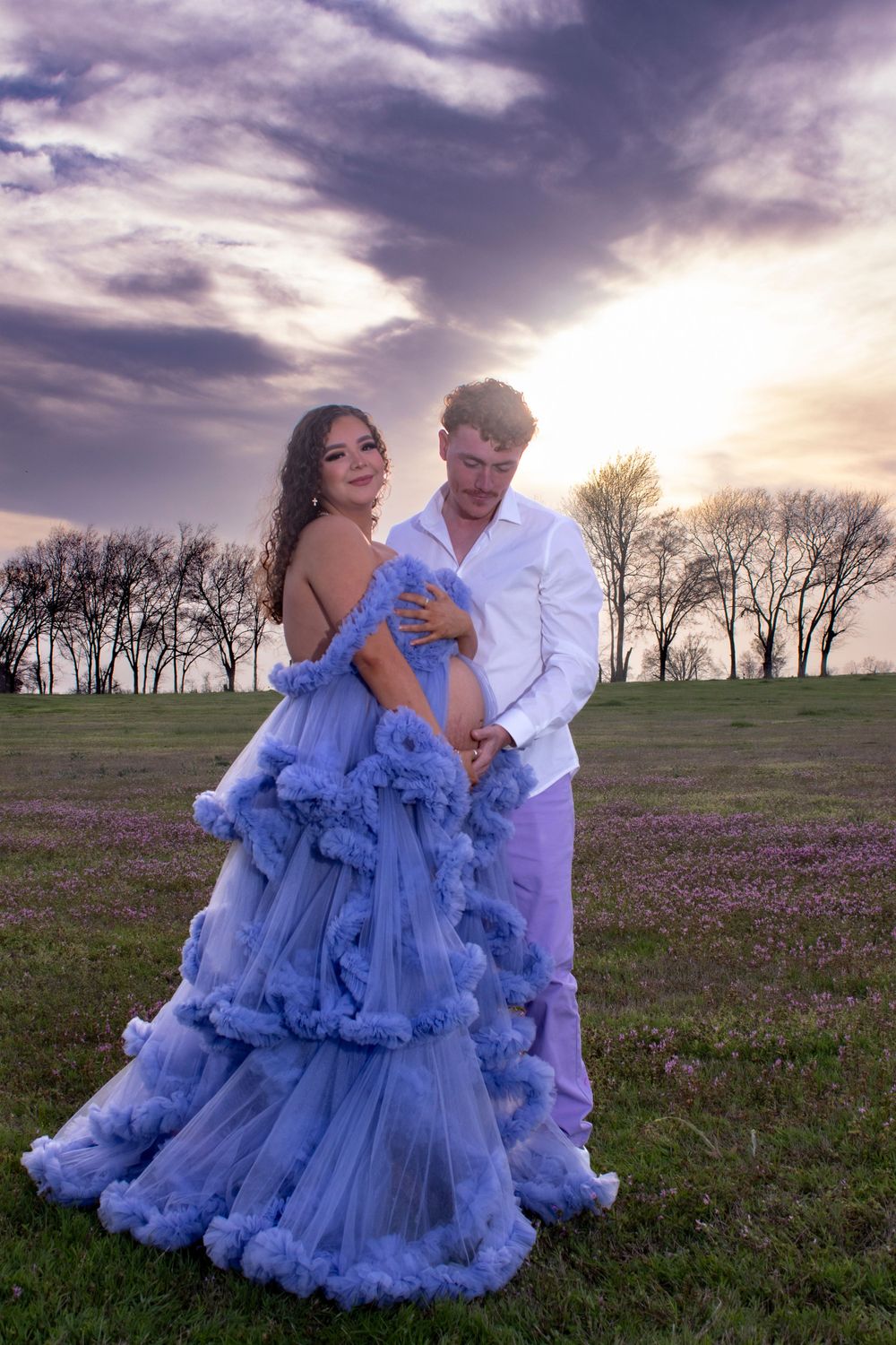 A couple embraces in a field of purple wildflowers at sunset, with the woman wearing a flowing blue tulle gown.