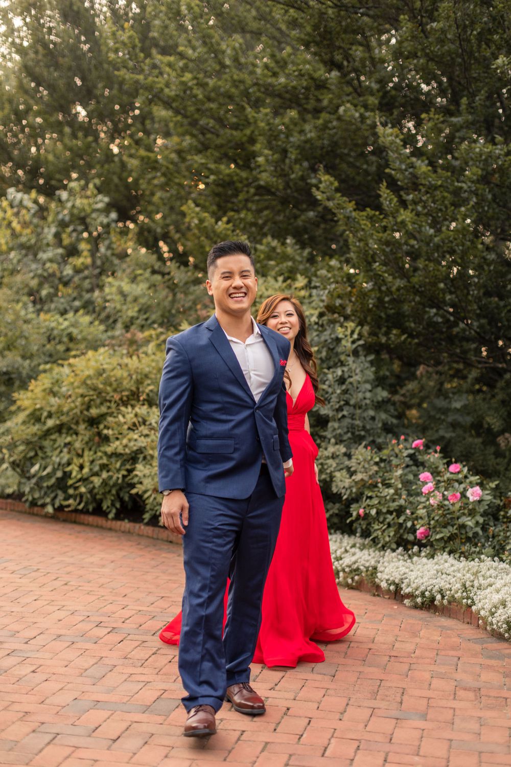 Couple in formal attire walks along brick pathway in garden setting.