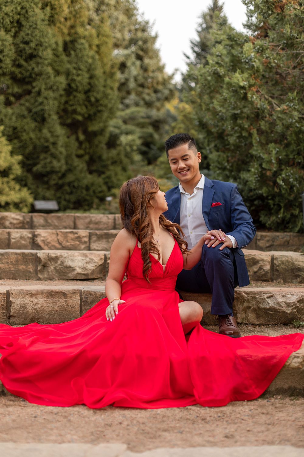 Elegant couple in formal wear pose on stone steps surrounded by evergreen trees.