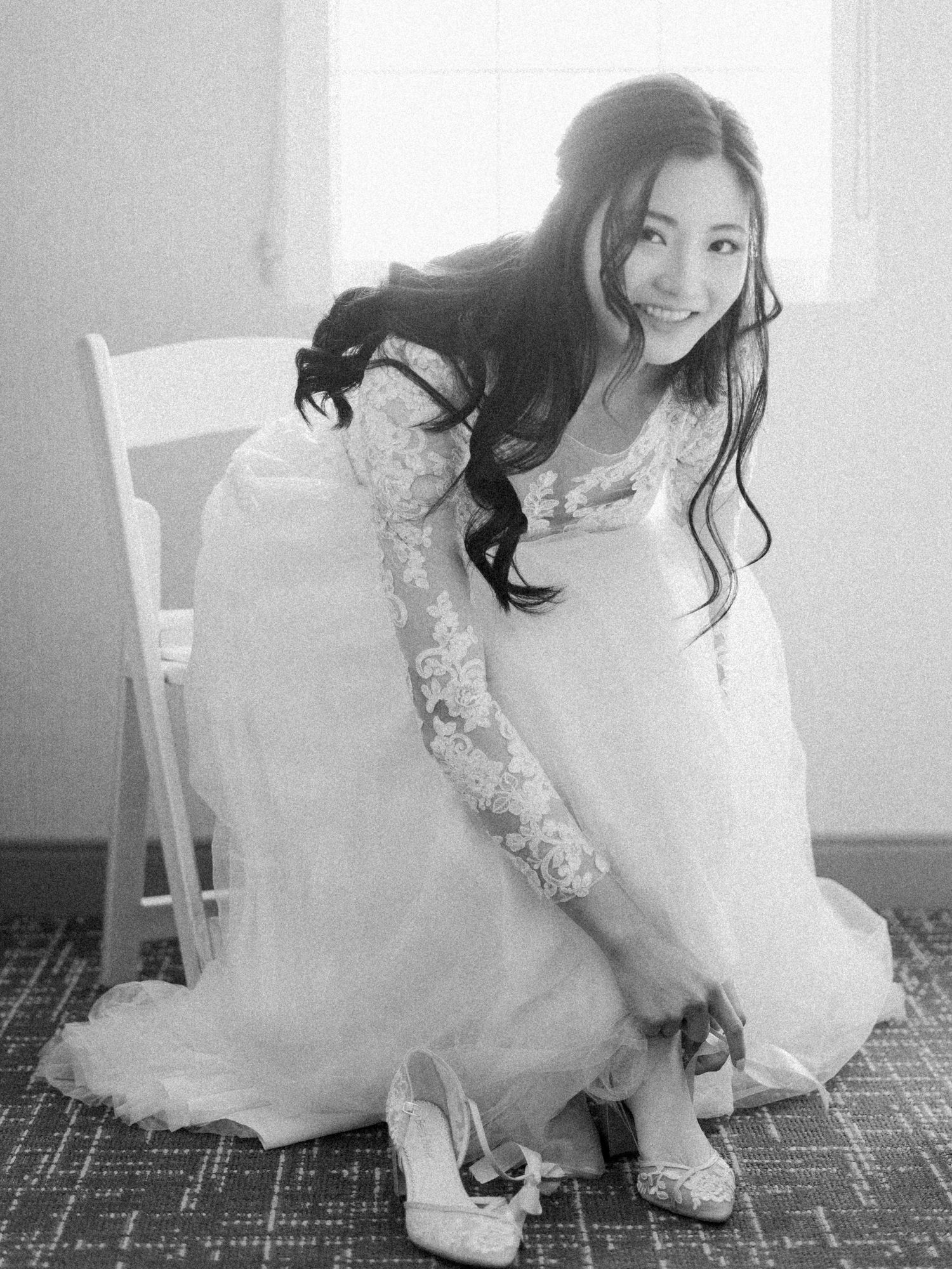 A bride adjusts her shoe while wearing a lace-sleeved wedding gown in an elegant black and white photograph.