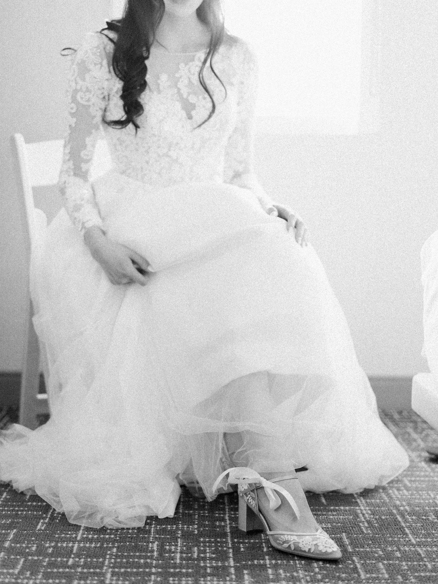 Black and white portrait of a bride seated in a white chair showing the details of her lace wedding dress.