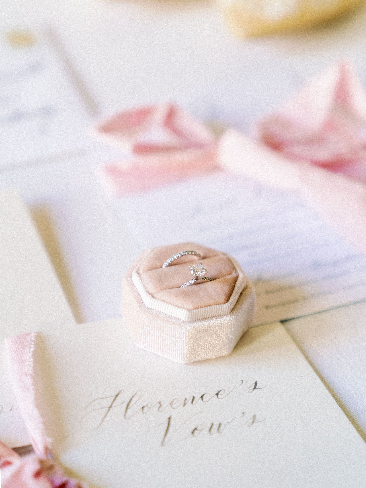 A close-up of an engagement ring in a blush velvet box with calligraphy and pink ribbons.