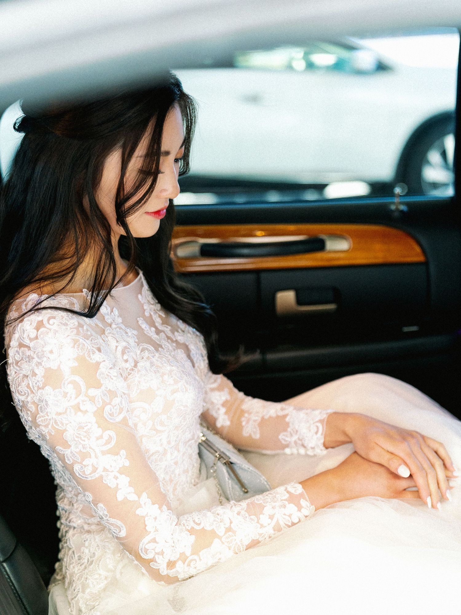 Bride in long-sleeved lace wedding dress sits in luxury car with wooden interior trim.