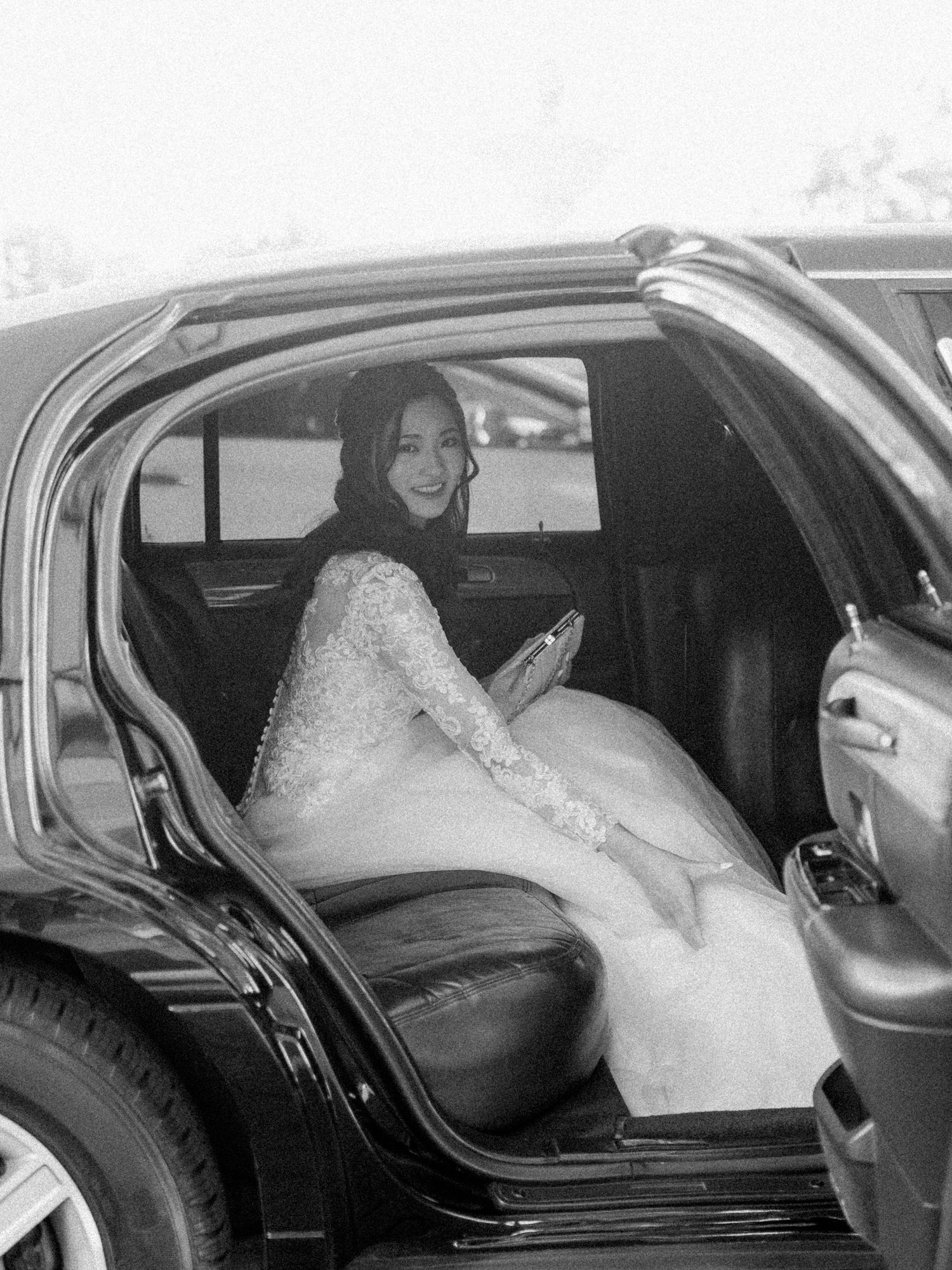 Black and white photo of a bride entering a classic car in her wedding gown.