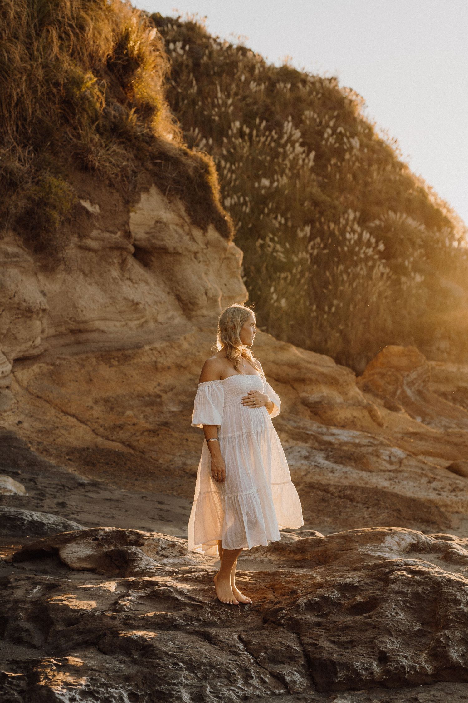 Woman in white off-shoulder dress poses on rocky coastal cliff at golden hour.