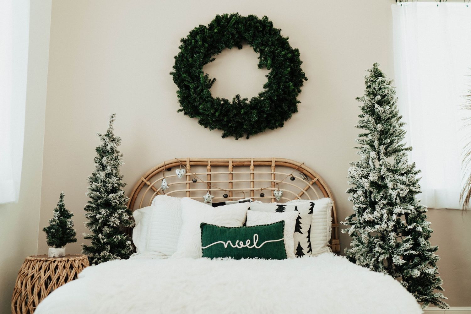 A cozy winter bedroom with two flocked Christmas trees, a rattan headboard, and a green wreath above the bed.