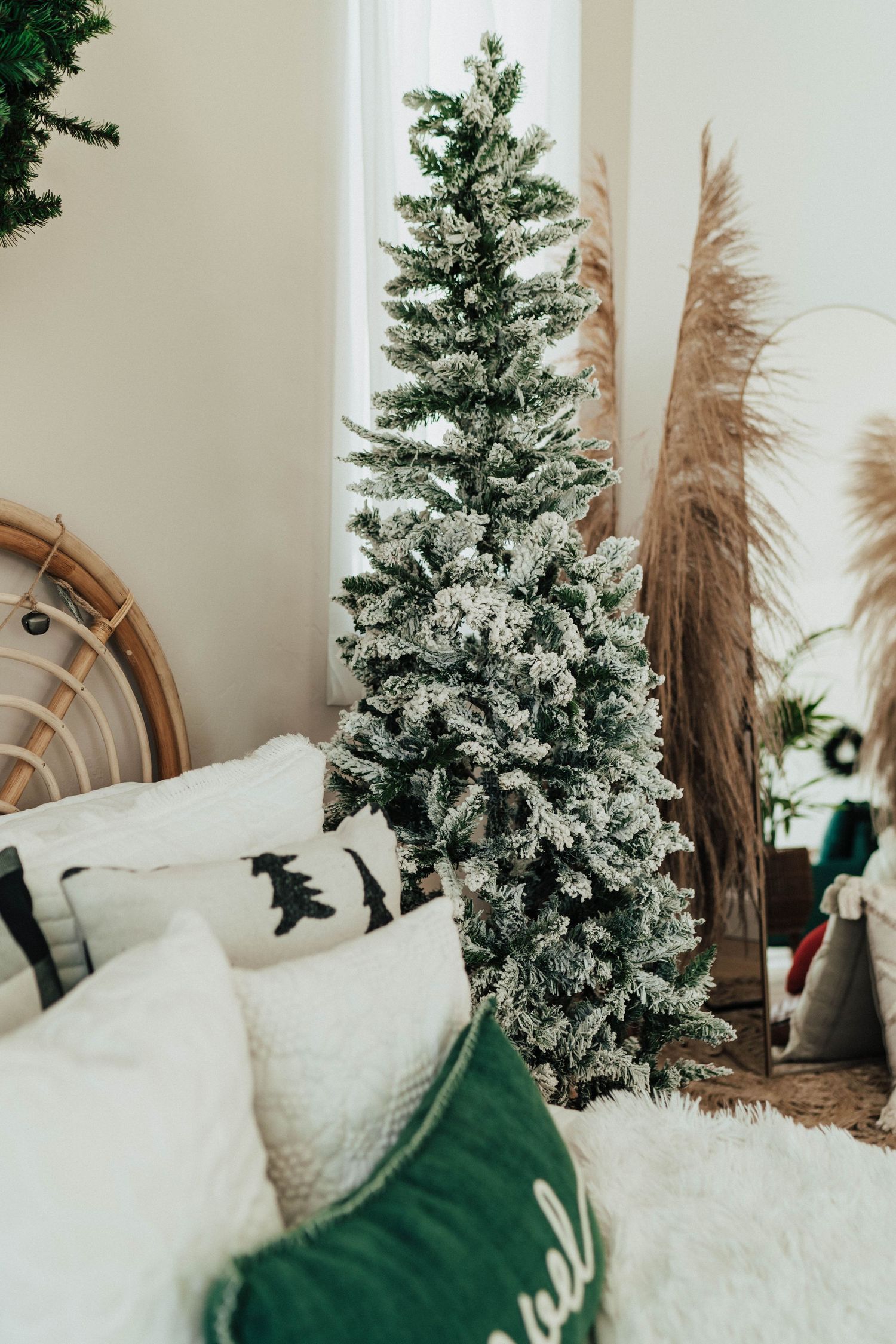 Close-up of a flocked Christmas tree with white bedding and a green pillow in a cozy bedroom corner.