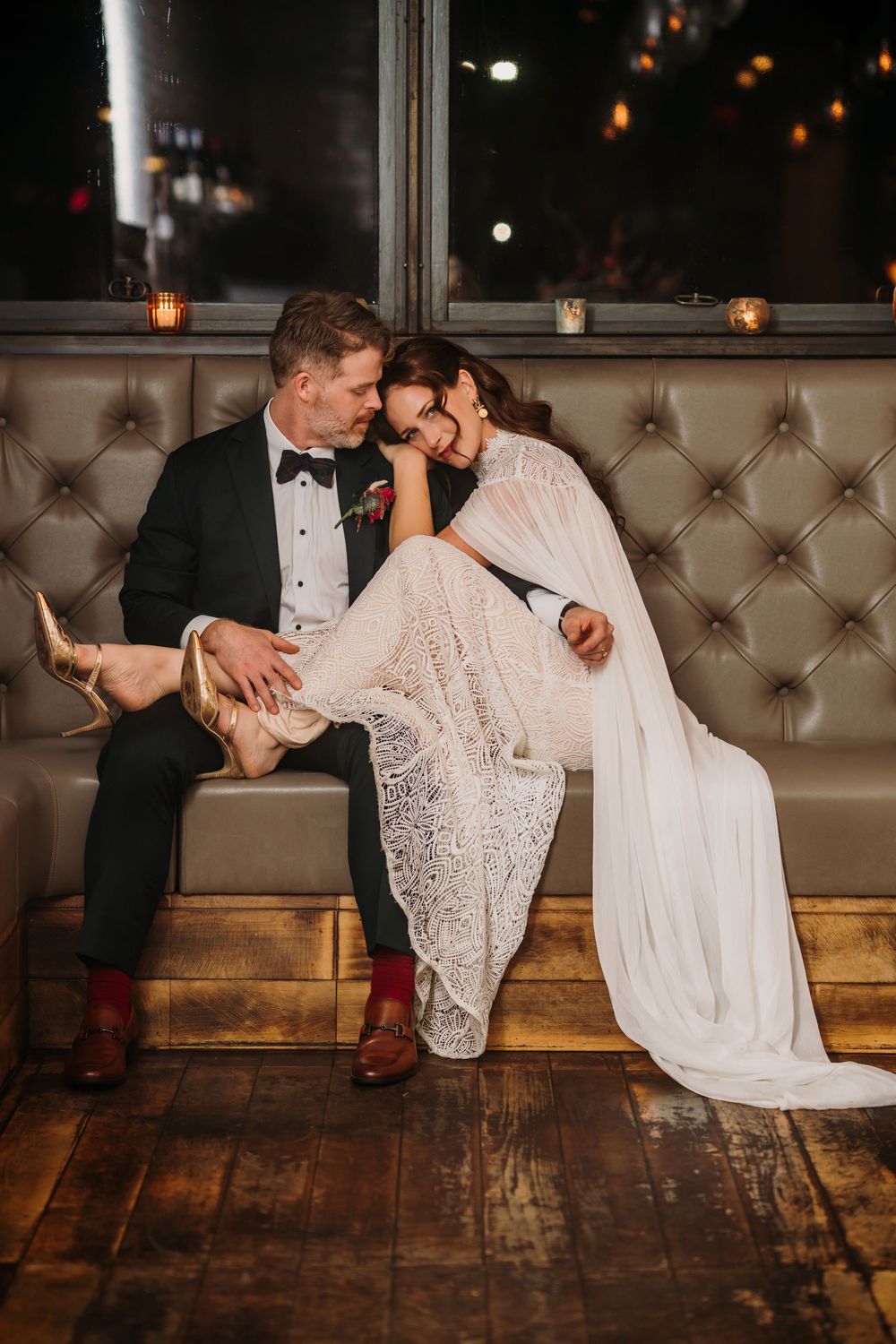 A romantic wedding couple shares an intimate moment on a tufted leather bench with candlelit ambiance.