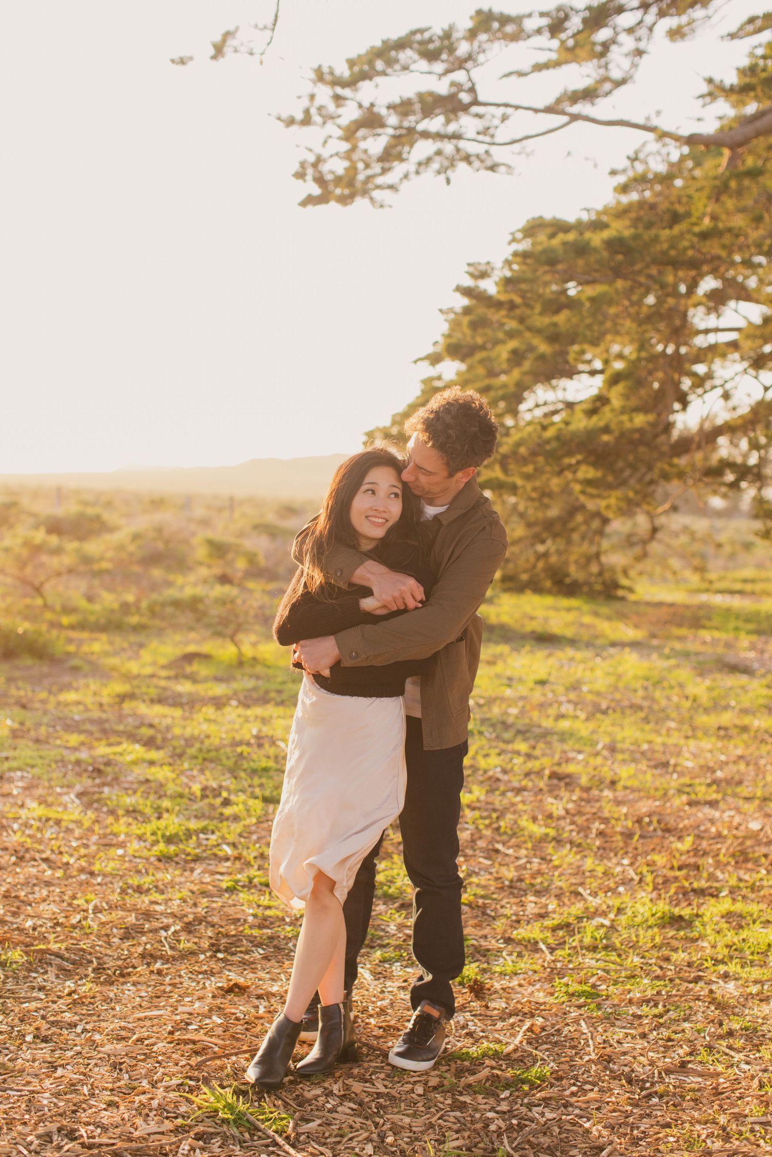 Romantic couple embracing in golden sunset light among tall trees in a park setting.
