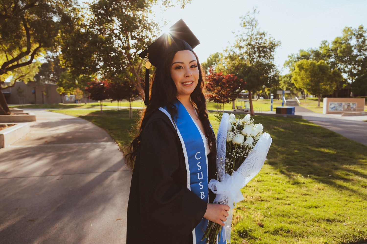 A graduate in a blue stole holds white flowers on a sunny campus walkway.