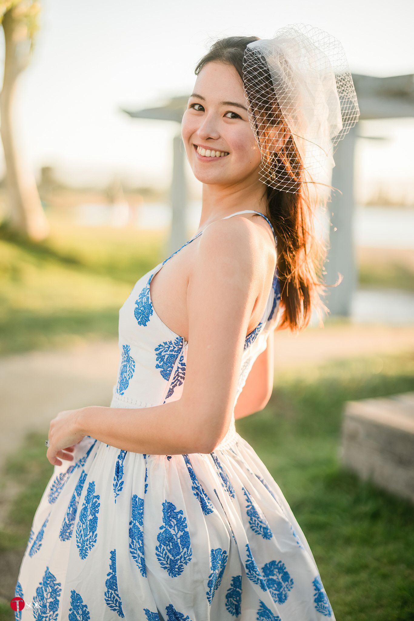 A woman wearing a veil and blue patterned dress stands in warm evening sunlight.