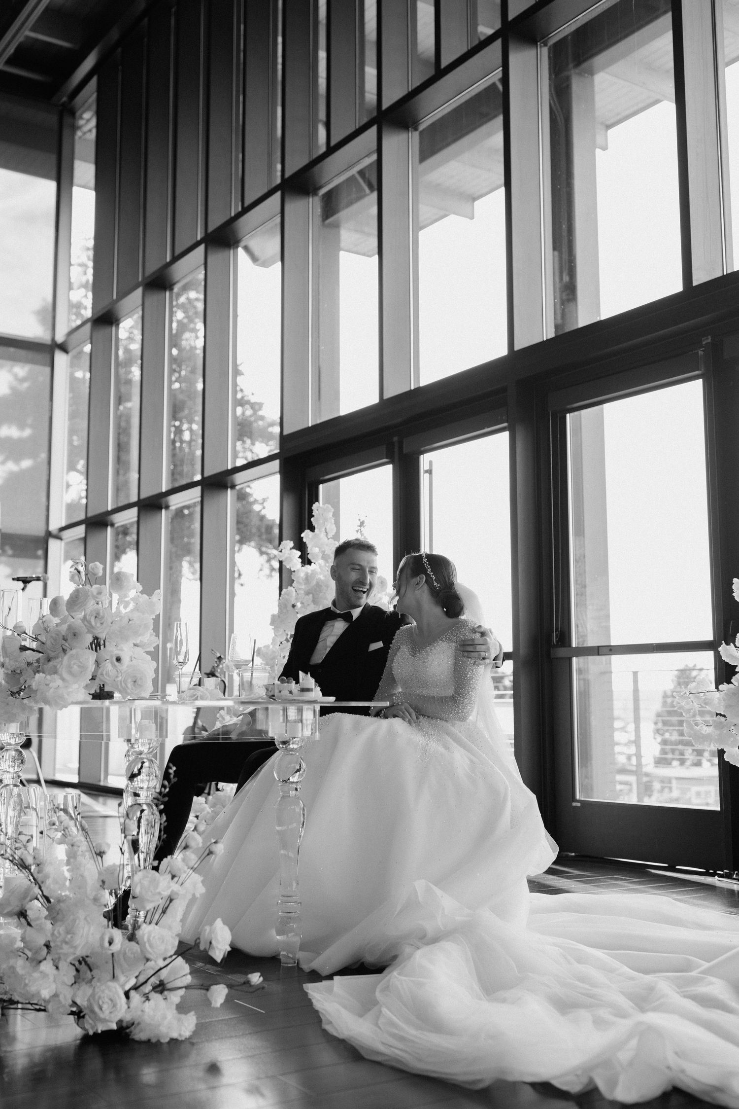 Black and white photo of wedding couple enjoying intimate moment at reception by large windows.