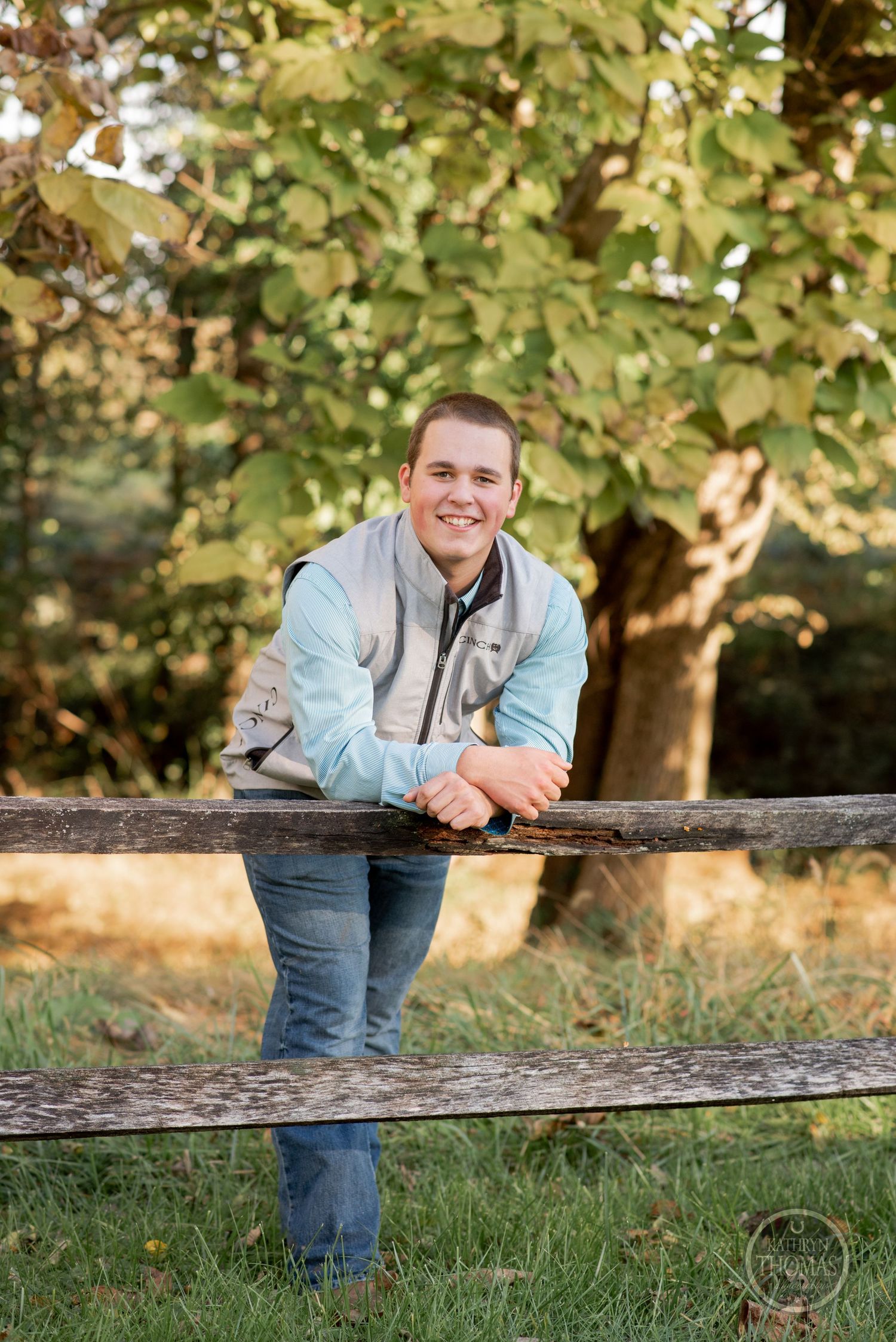 Person in athletic vest leans on wooden fence with autumn foliage in background.