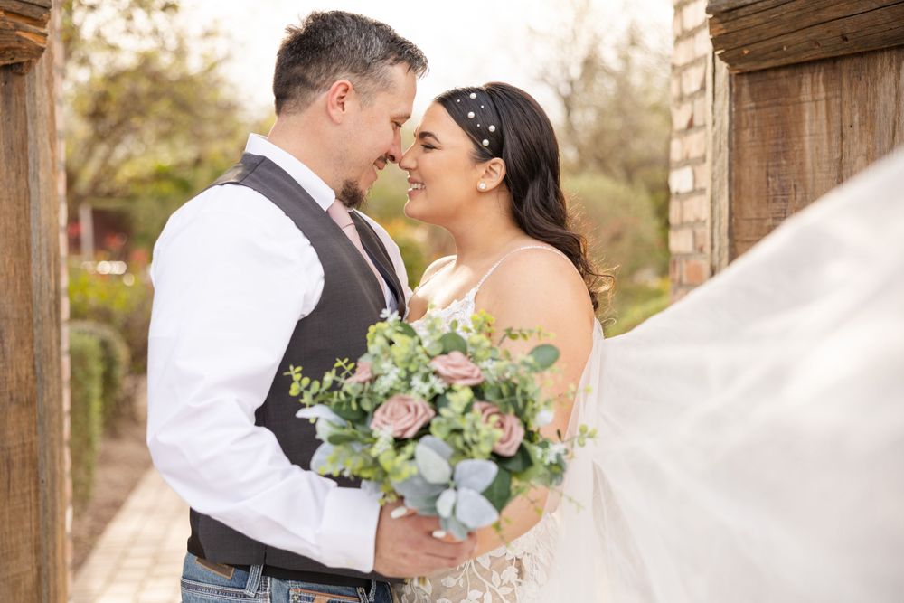 Romantic wedding portrait under wooden archway with soft pink floral bouquet.