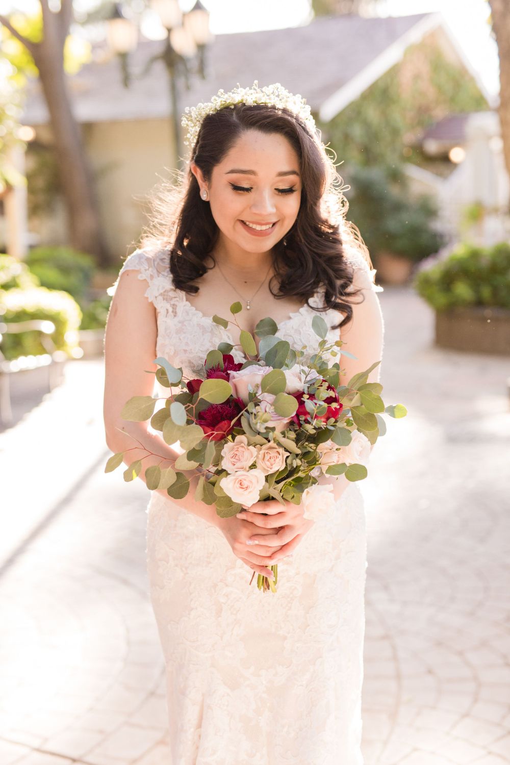 Bride in lace gown holding romantic bouquet with red roses and eucalyptus in sunlit garden.