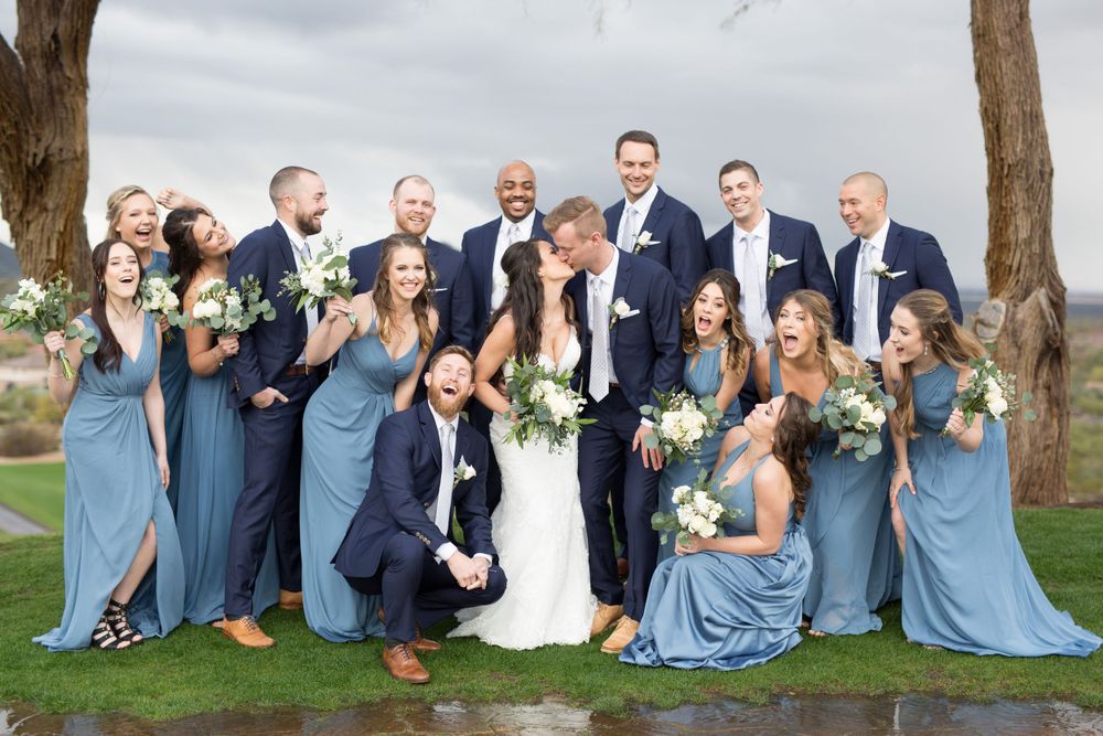Wedding party in blue dresses and navy suits poses together under trees.
