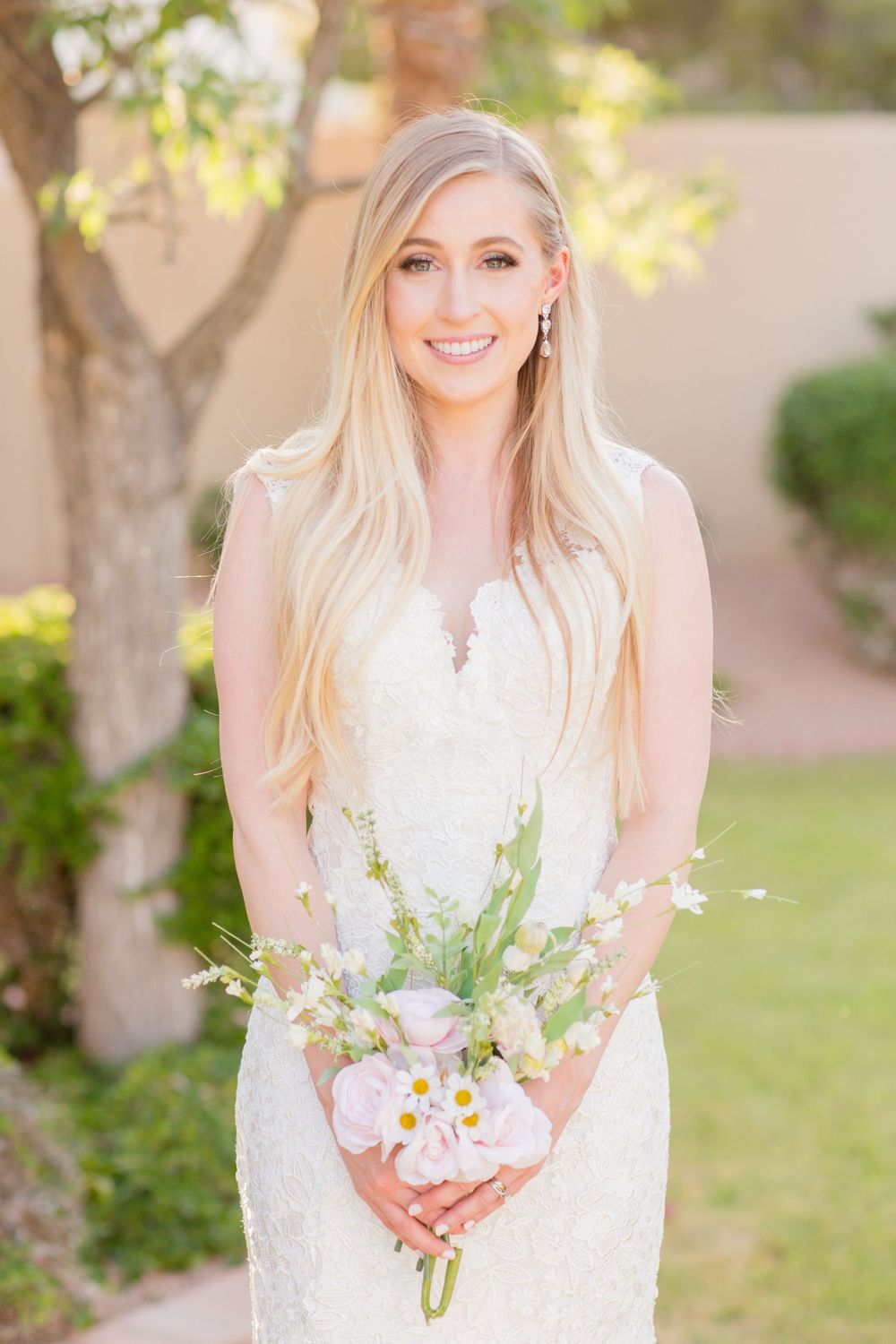 A bride in a fitted lace gown holds a delicate pink and white bouquet in a garden setting.