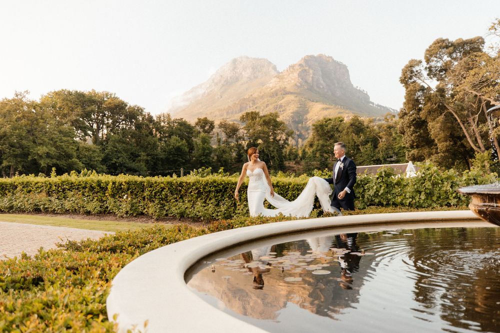 bride leading the way while groom carries her train in front of a fountain at Molenvliet