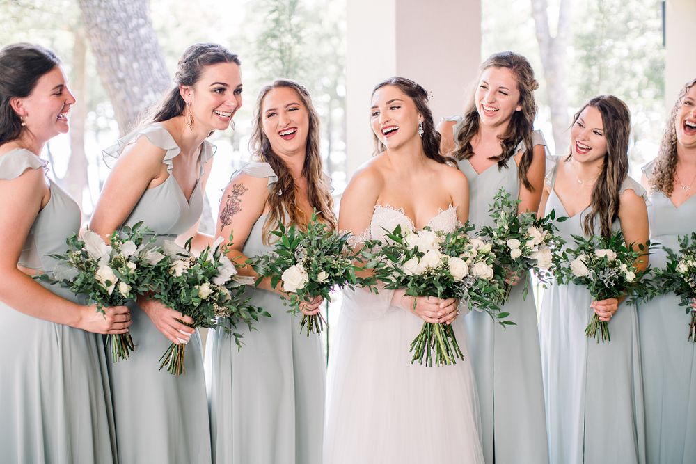 Bridesmaids in light blue dresses laughing while holding white and green floral bouquets.