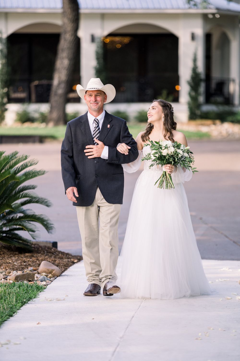 Father in cowboy hat escorts bride in flowing white gown down garden path.