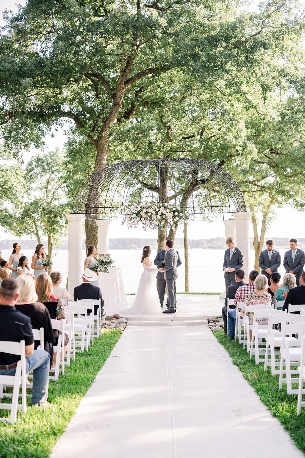 Wedding ceremony setup with white chairs lining a pathway under tall trees in an outdoor venue.