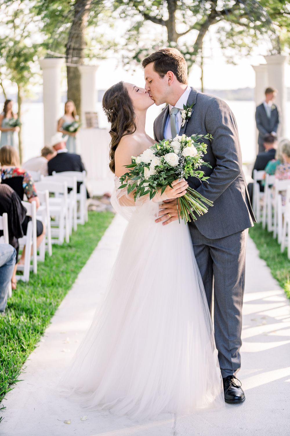 Newlyweds share a romantic moment on a paved pathway lined with white chairs outdoors.