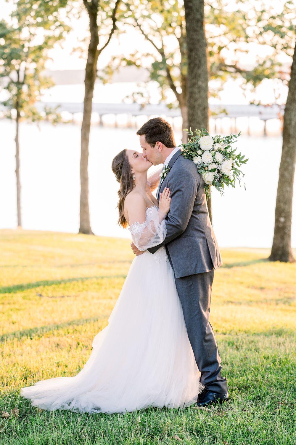 Tender moment between newlyweds on grassy lawn with trees and water in background.