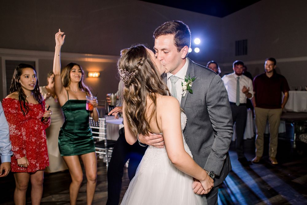 Couple sharing a dance at their wedding reception while guests in red and green dresses dance in the background.