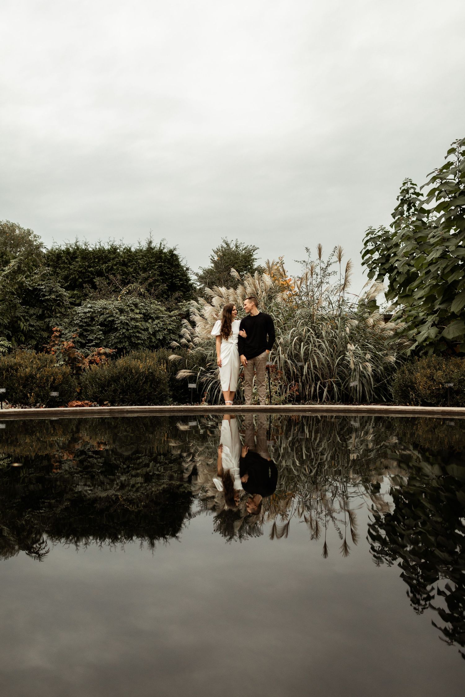 Artistic reflection shot of couple standing by still water in garden setting.