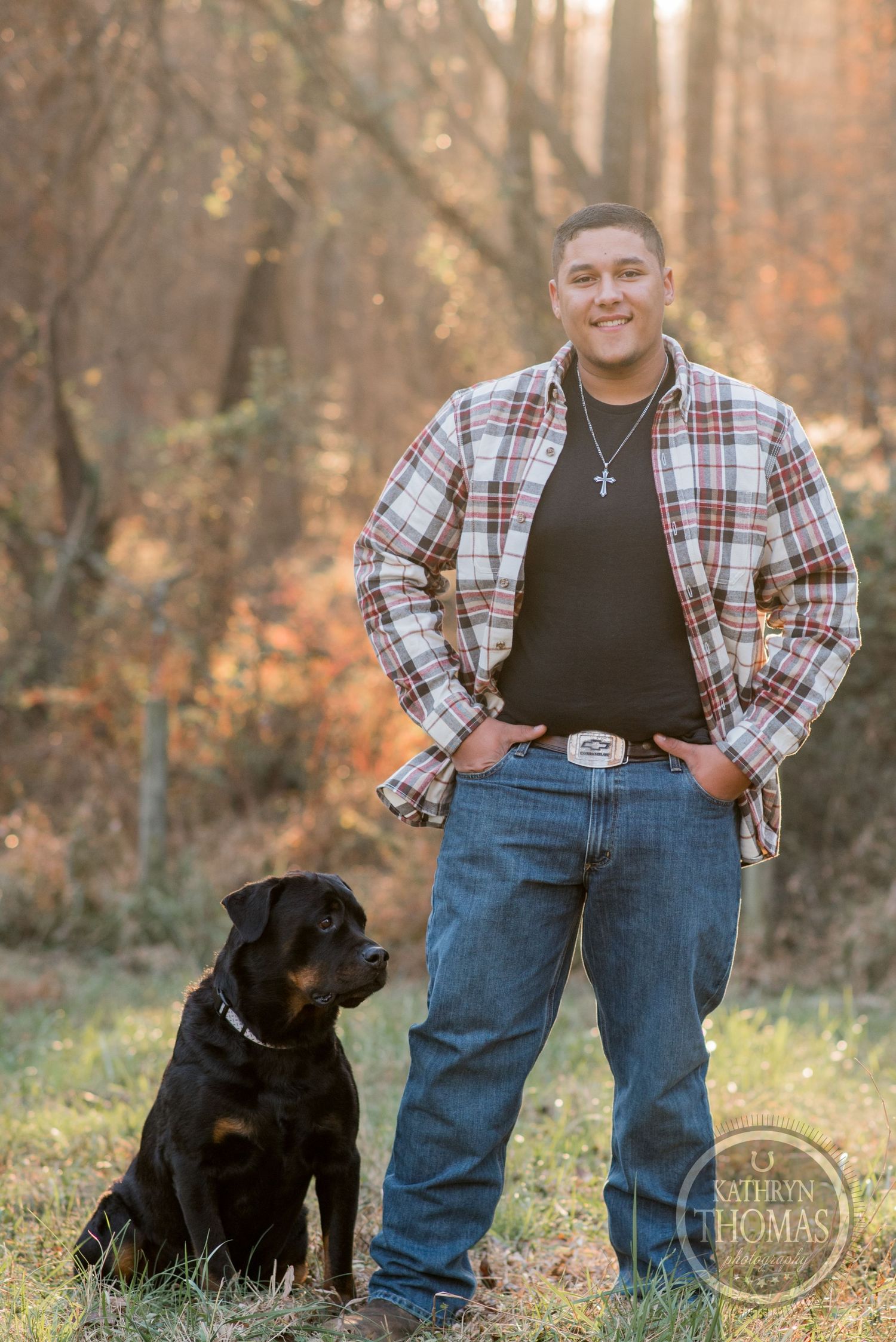 A black Labrador sits beside someone wearing plaid and jeans in an autumn forest setting.