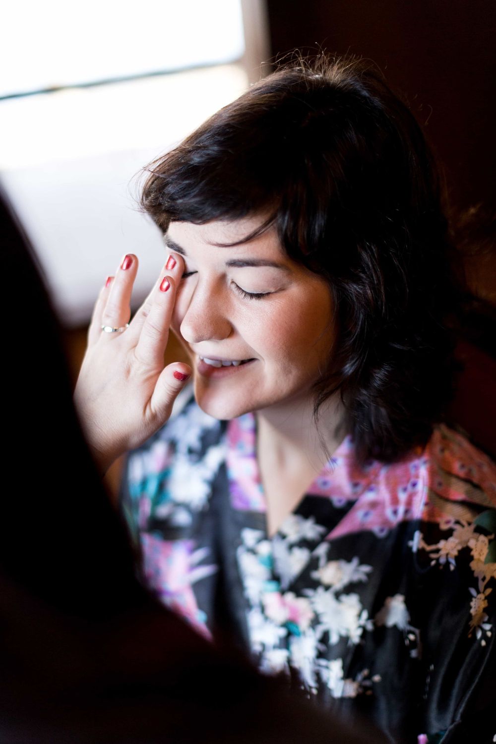 Dark-haired bride in floral robe touching her face while getting ready on wedding day.