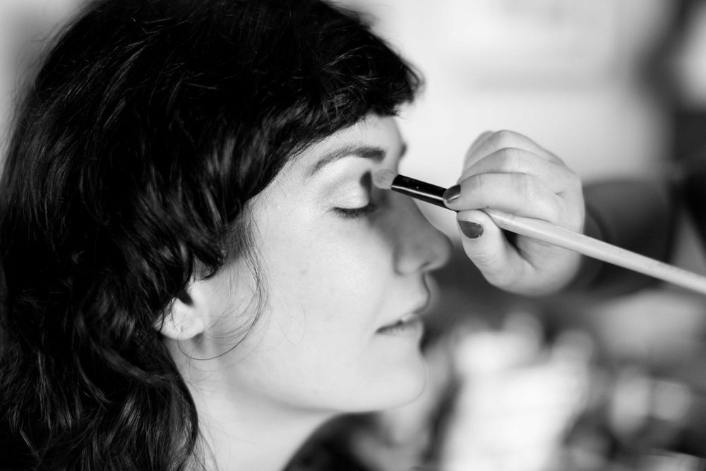 Black and white photo of bride getting eye makeup applied with a brush before wedding.