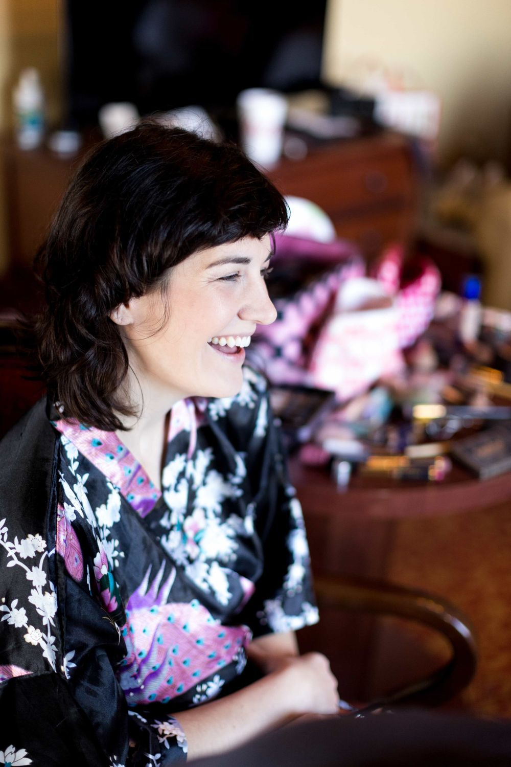 Laughing bride in black floral robe sitting in chair during wedding day preparations.